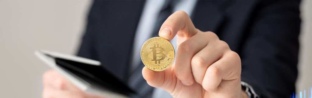 Businessperson holding a Bitcoin coin in front of a tablet, representing digital currency transactions, crypto investment, and blockchain-based financial technology.