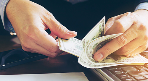 Businessperson counting dollar bills near a laptop, symbolizing financial management, cash flow analysis, and investment planning in corporate finance.