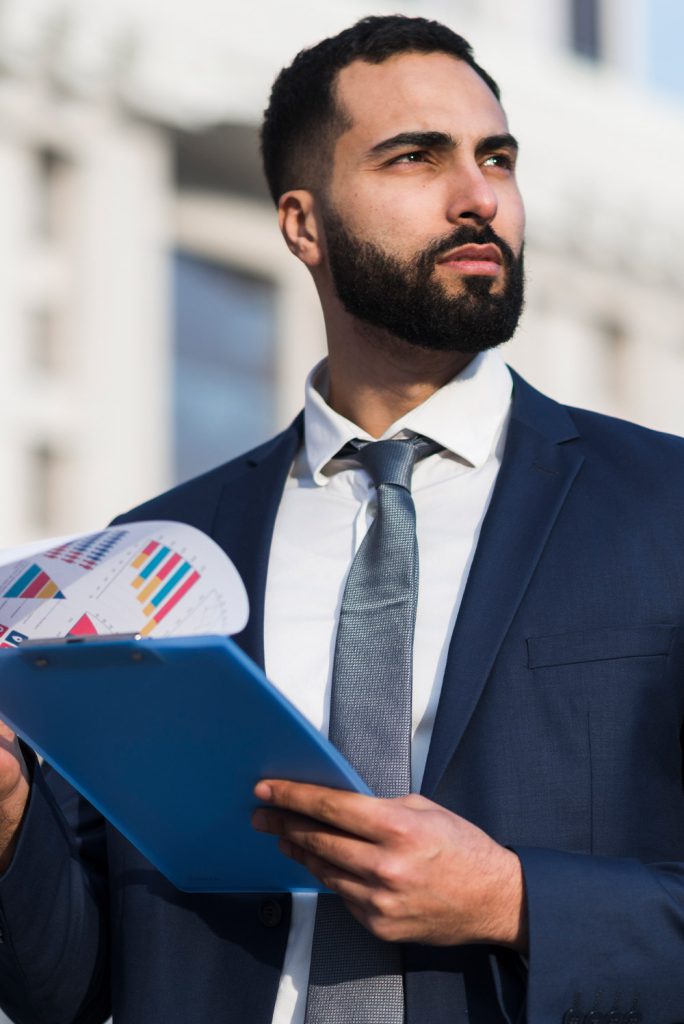 Confident businessman holding a blue clipboard and financial charts while looking ahead outside a modern office building.