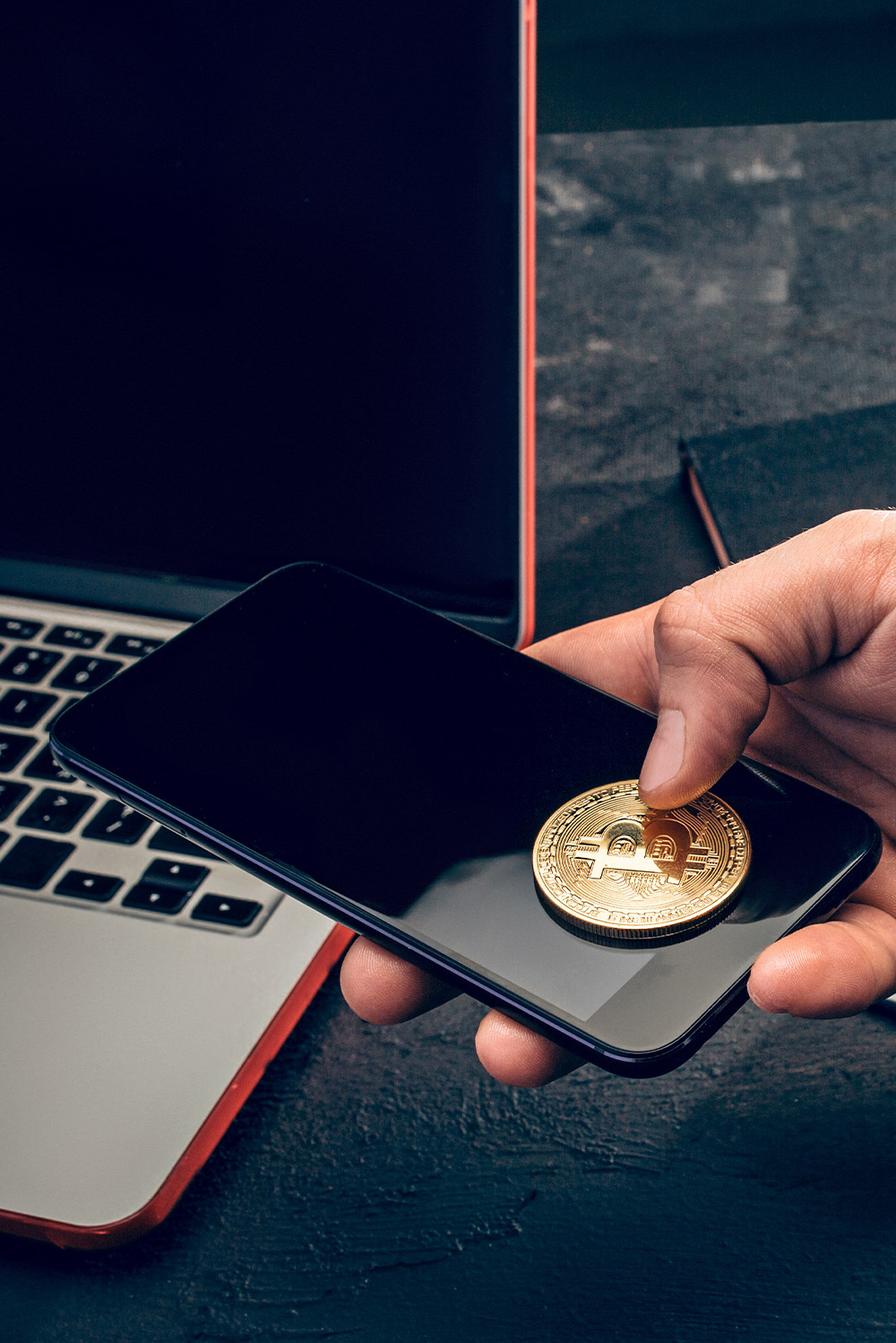 Hand placing a Bitcoin coin on a smartphone near a laptop, symbolizing mobile cryptocurrency transactions, digital wallets, and modern blockchain technology.