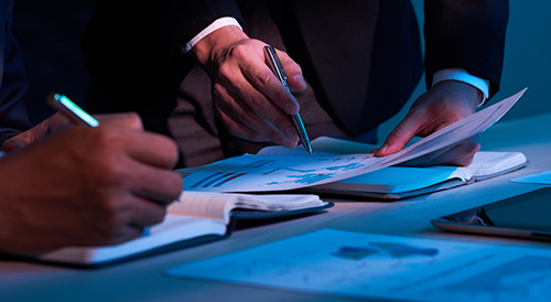 Business professionals reviewing financial documents and taking notes during a meeting at a blue-lit workspace.