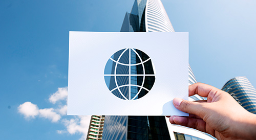 Hand holding a paper cutout of a globe icon in front of modern glass skyscrapers, symbolizing global reach and international business.
