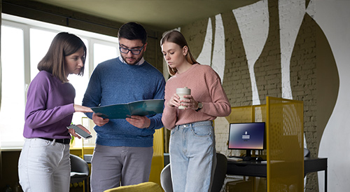 Three young professionals standing in an office discussing a project, reviewing documents and using a tablet, symbolizing teamwork, business planning, and collaborative strategy.