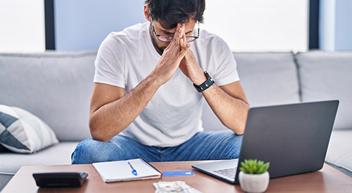 Stressed young man sitting on a couch with a laptop and financial documents, representing financial risk, debt management, or startup budget challenges.