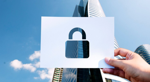 Hands holding a paper cutout of a padlock against a background of modern skyscrapers, representing data protection, cybersecurity, and secure digital infrastructure in real estate or technology sectors.