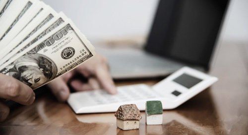 Close-up of a person holding US dollar bills beside miniature house models and documents on a desk, symbolizing real estate investment, property financing, and financial planning.