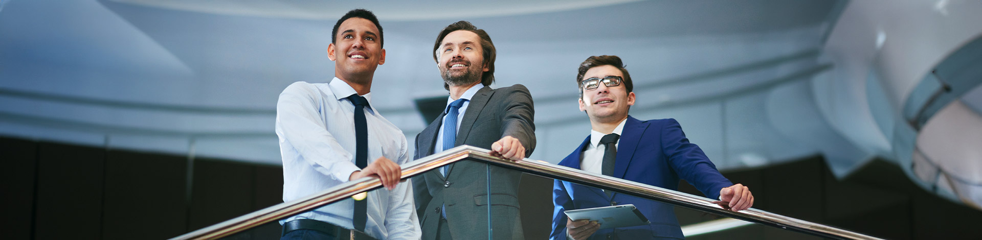 Three confident business professionals standing on a staircase railing, looking forward in a modern corporate building.