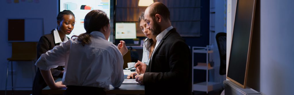 Business team discussing documents during a late-evening meeting, with diverse colleagues collaborating around a table in a modern office.